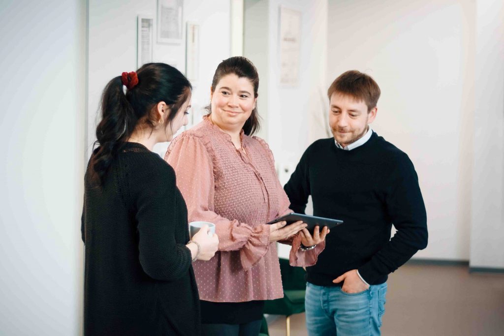 Drei Personen, zwei Frauen und ein Mann, stehen im Gespräch zusammen, wobei einer ein Tablet hält, in einem hellen Büroumfeld.